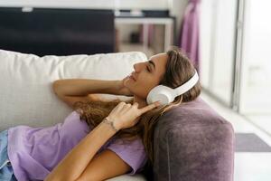 Smiling woman listening to lively music by big white earphones. Enjoying time at home , lieing on sofa. photo