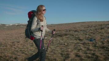 Smiling woman in sunglasses with backpack and trekking poles is hiking video