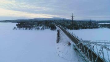 Car Goes on Winter Road at Sunset. Bridge over the Frozen Lake. Aerial View. Drone Flying Backward, Camera Tilts Down. Establishing Shot.. video