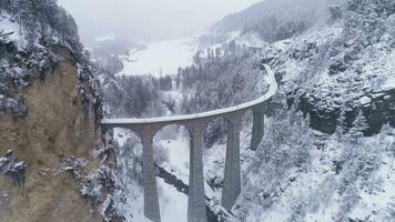 terraplén viaducto con ferrocarril a invierno Nevado día en Suiza. aéreo vista. suizo Alpes. nevando zumbido moscas adelante, cámara se inclina arriba video