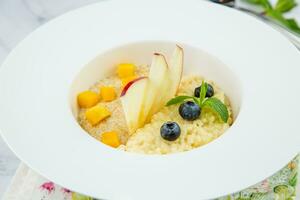 porridge with pieces of apple, mango, berries and mint in a white plate side view photo