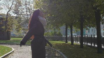 Young beautiful caucasian brunette southern smiling woman with curled hairs in grey coat and white scarf is walking towards the camera and turning around in park. Slow motion. Steadicam medium shot. video