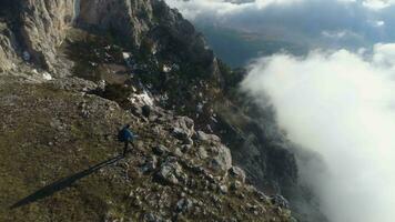 escursionista uomo con zaino è a piedi e in piedi su il ripido bordo di montagna altopiano sopra il nuvole. aereo Visualizza. fuco è orbitante. video