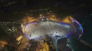 A crowd of people are skating on large illuminated ice rink in the evening. Christmas time. Aerial view. Camera is tilting down. Establishing shot. video