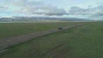 Suv Car Goes on Gravel Road in Summer Day. Mountains and Lake Shore on Background. Aerial View. Drone is Orbiting around. Establishing Shot video