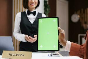 Adult with greenscreen at front desk, talking to reception staff about hotel room service and holding device. Guest looking at chroma key display with isolated mockup template in lobby. photo