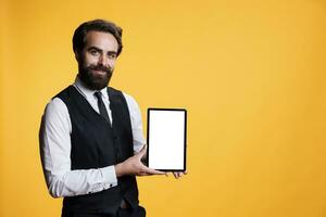 Smiling worker presents white display on tablet, posing with confidence against yellow background. Young man butler showing blank empty copyspace screen in front of camera, mockup template. photo