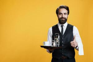 Happy waiter does thumbs up sign while he holds platter with coffee and accessories in studio, professional stylish butler. Man restaurant employee showing like agreement gesture on camera. photo