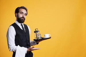 Skilled elegant butler pointing aside and holding food platter to serve customers, working at five star restaurant. Young adult dressed in suit and tie carries tray with cup of coffee. photo