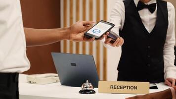 Receptionist using POS payment terminal to validate card purchase of room services and additional amenities, close up. African american guest using smartphone banking app during check in process photo