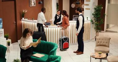 Focused hotel manager verifying tourists booking paperwork while they are being assisted by concierge personnel. Bellboy helping guests with baggage after check in process is over. photo