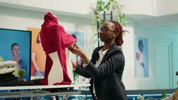 méticuleux vente au détail assistant travail dans luxe mode boutique, en mettant sur femmes chemisier sur mannequin. expérimenté africain américain employé mettant en valeur Nouveau collection de branché habits video