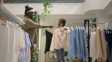 Pareja en ahorro tienda mira a ropa, molesto a encontrar barato pero elegante formal atuendo para su boda. mujer Ayudar marido escoger Entre elegante chaquetas americanas en segundo mano ropa Tienda video