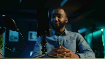 Close up of cellphone on holder and microphone used by online star in dimly lit home studio to film video for online platforms. Phone being used by african american influencer to record footage