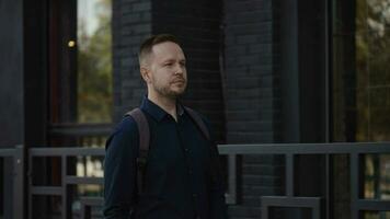 A young man in a good mood walks along a city street. Portrait of a city guy in a shirt and with a backpack behind his back video