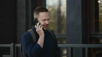 een aantrekkelijk en elegant stedelijk Mens is wandelen naar beneden de straat en pratend Aan de telefoon. een mooi zo humeur en glimlach begeleiden een stad bewoner. video