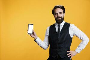 Professional waiter points at white screen on smartphone, presenting isolated copyspace template on display in studio. Experienced butler presents empty mockup modern device on camera. photo