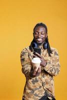 African american man taking photo of cup with coffee using mobile phone, enjoying free time in studio over yellow background. Cheerful smiling person drinking beverage, listening music and having fun