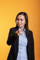 Filipino woman in formal suit pointing at camera with fingers. Portrait of beautiful cheerful young adult with casual attitude using charisma, standing in studio over yellow background photo