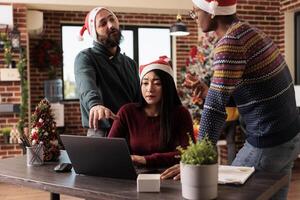 Diverse men and woman coworkers brainstorming while working on project together on xmas eve. Company employees team in santa hats discussing task on laptop on christmas in office photo