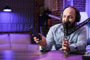 Portrait of tech vlogger reviewing fresh mobile phone release in rgb lights home studio. Entertaining content creator filming technology channel episode for interested audience photo