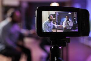 Close up shot of professional camera used to record podcast show with vlogger and guest discussing in blurry background. Equipment in living room personal studio used for live streaming broadcast photo