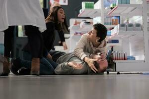 People holding man with seizure symptoms, screaming for help in medical retail store. Drugstore client fainted near medication shelf and having epilepsy disorder attack on pharmacy floor photo