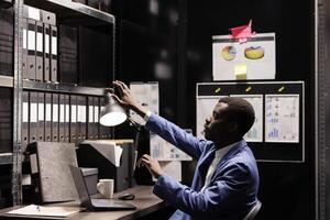 African american inspector enjoying using 3d simulation to solve missing person case, searching for new evidence using innovative technology. Police officer working overtime in arhive room. photo