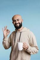 Cheerful arab man holding coffee cup, smiling and showing ok gesture with fingers studio portrait. Carefree person drinking hot tea from white mug and looking at camera with approval sign photo