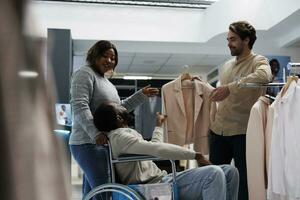 Man with physcial disability choosing jacket with girlfriend while shopping in clothing store. Department mall african american customer in wheelchair selecting outfit with wife photo