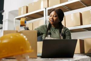 almacén supervisor trabajando a clientela pedidos preparando paquetes para entrega, comprobación Envío detalles en ordenador portátil computadora. africano americano empleado analizando bienes calidad controlar reporte foto