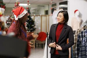 Asian client trying on blazer before buying festive clothes, employee helping woman to choose outfit for christmas dinner with family. African american assistant recommending suit to client. photo