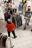 Various shoppers looking at clothing items on racks in shopping center while seeking for garments to purchase. Consumers going apparel shopping as gifts for festive season, Christmas eve. photo