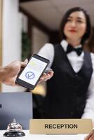 Man paying with mobile phone and nfc at pos terminal, receptionist helping people with check in process. Asian front desk staff welcoming guests, adult making payment for room. Close up. photo