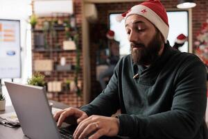Startup company worker typing message on laptop while working in office at christmas eve. Corporate employee wearing santa claus hat using computer and sitting at workplace desk photo