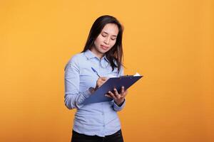 Cheerful filipino woman writing business report on files, analyzing questionnaire and answering question in studio. Confident adult standing over yellow background looking at checklist photo