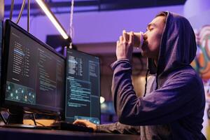 Hacker breaking website on computer while drinking coffee to go. Young asian man in hood cracking password using malicious software and getting illegal access to steal data photo
