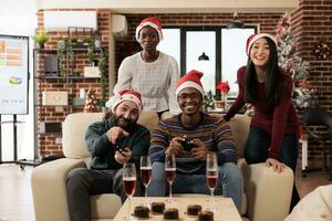 puesta en marcha diverso empleados equipo disfrutando festivo Navidad celebracion en oficina. colegas Bebiendo y jugando vídeo juegos con palancas de mando a invierno estacional corporativo fiesta foto