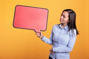 Filipino woman holding empty red speech bubble with copy space, advertising product. Cheerful young adult standing in studio over yellow background, looking at blank dialog frame with place for text photo