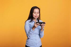 Playful gamer playing video games on mobile phone, trying to win gaming competition. Filipino woman having fun with online gameplay as leisure activity, standing in studio over yellow background photo