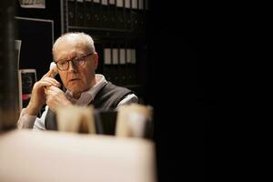 Elderly investigator discussing criminal case with remote officer using landline phone, working late at night in arhive room. Private detective analyzing criminology report. Investigation concept photo