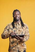 African american man holding tablet computer, browing on internet in studio over yellow background. Smiling adult scrolling trough social media, watching funny video on digital device photo