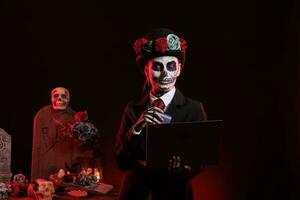 Young adult doing online shopping with skull make up, using credit card on laptop to make payment. Wearing santa muerte lady of death costume to celebrate mexican halloween holiday. photo