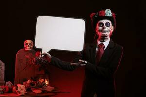 Woman dressed in la cavalera catrina holding speech bubble with carnival make up and traditional day of the dead costume. Advertising isolated mockup cardboard and looking like santa muerte. photo