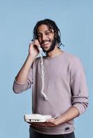 Happy arab man smiling while answering landline phone portrait. Cheerful handsome call center worker in casual clothes talking on telephone and laughing while looking at camera photo