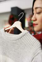 Client looking at clothing on hangers before buying presents for christmas eve winter festivity, person checking sweater material. Asian woman customer checking items on sale in store. Close up. photo