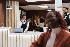 Employee making online room booking on phone, talking on landline call to note room reservation in hotel records. Female reception worker helping future resort guests with accommodation. photo