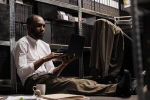 Private detective conducting investigation using laptop while sitting on floor surrounded by police report papers. Detective analyzing crime case information on computer in evening photo