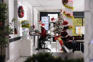 Multiethnical business team wearing christmas hats doing project tasks in festive workplace. Multicultural employees checking statistical graphs in decorated office during xmas season photo