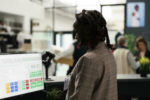 Showroom employee analyzing merchandise inventory on computer, working at clients purchase report in shopping centre. African american worker standing at cash register in modern boutique photo